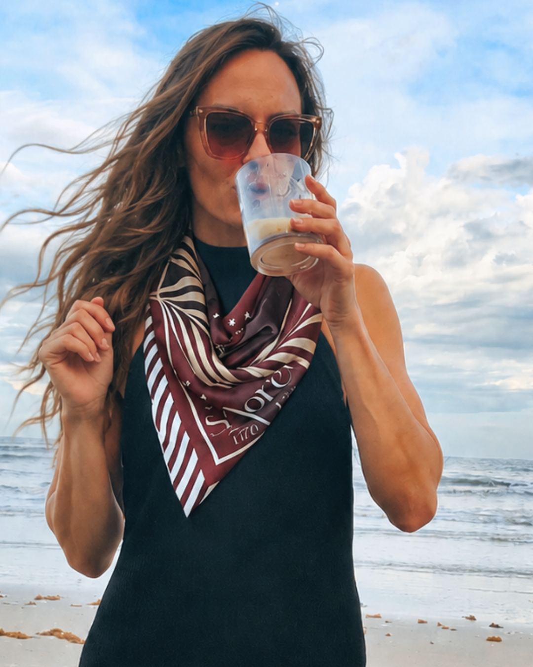 Woman drinking from a glass on a beach with a scarf around her neck